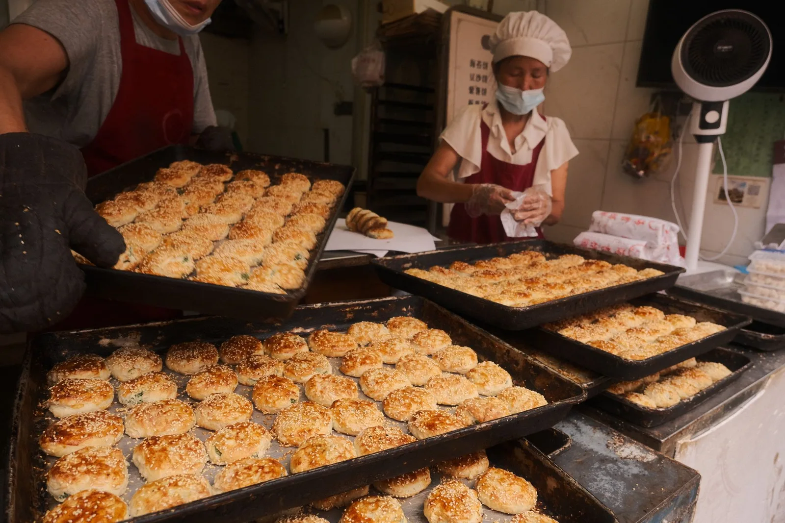 Bakery staff pulling trays of fresh sesame pastries from a busy production line