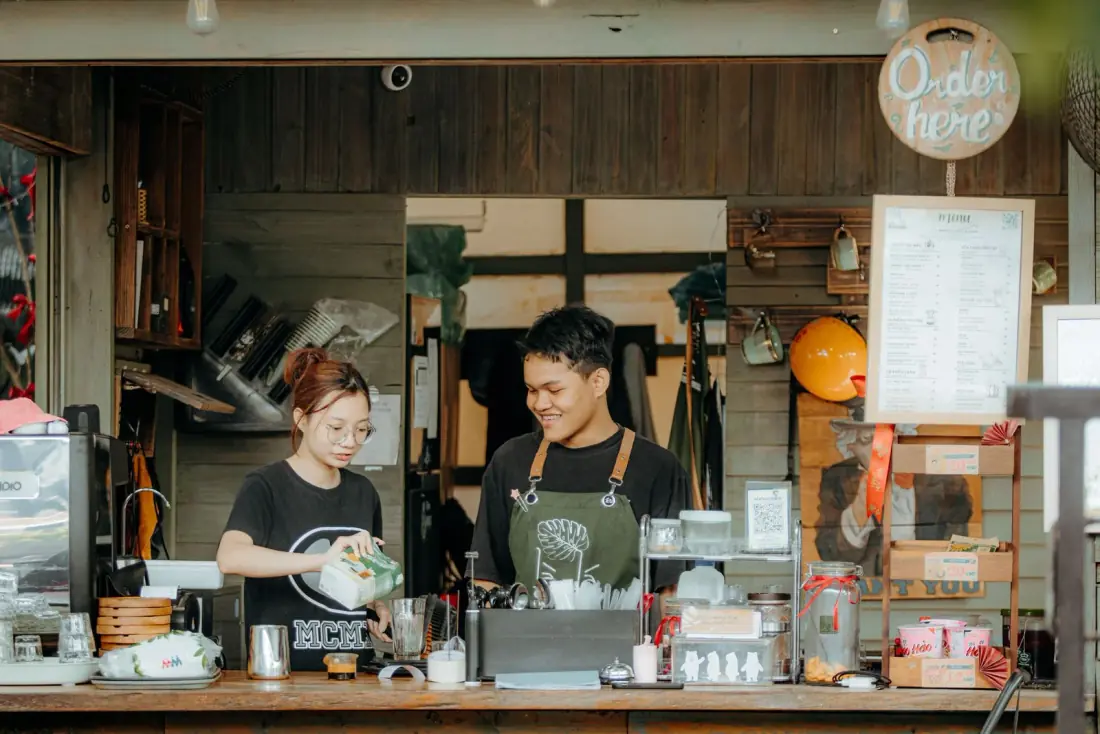 Two young staff members serve customers behind a busy café counter beneath an 'Order here' sign.
